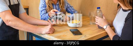 Unrecognizable woman making contactless credit card over a payment terminal held by waiter at cafe Stock Photo