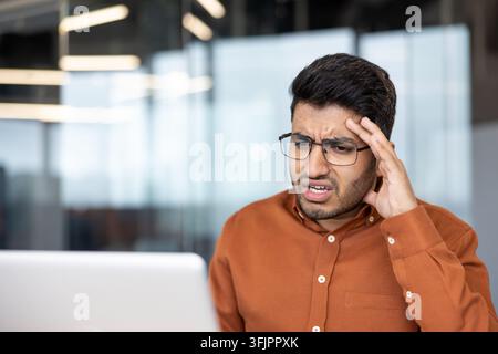 Burnout and workplace stress, young businessman looking at laptop with frustration and headache while working in modern office environment during a busy workday. Professional employee feeling pressure Stock Photo