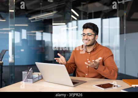 Businessman having a video call on a laptop in a modern office, smiling professional man using technology for remote communication and virtual meeting at his desk with documents and tablet. Stock Photo