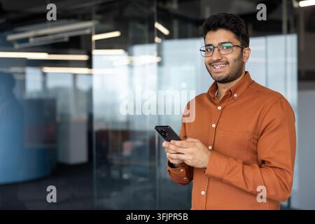 Businessman using smartphone in modern office, young professional man holding phone for digital communication and mobile technology tasks in a corporate workplace environment with glass partitions. Stock Photo