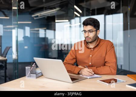 Businessman working on laptop and writing in notebook at office desk, professional man analyzing financial data and documents during a corporate meeting in modern workspace with digital technology. Stock Photo