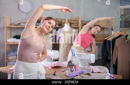 Two female dressmakers doing a warm-up for the muscles. Seamstresses rest and warm up during a break, they stand in the workshop against the backgroun Stock Photo