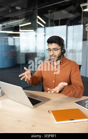 Customer support representative using a laptop and headset for a video call in a modern office. Professional man talking during a virtual meeting with hand gestures to explain business concepts. Stock Photo