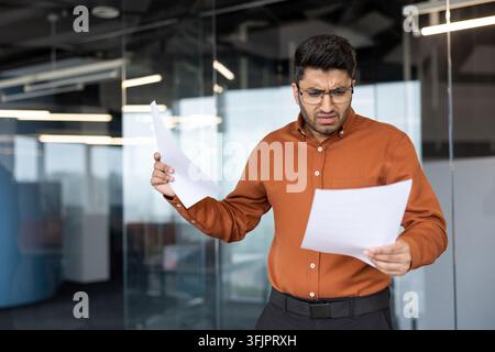 Confused businessman holding documents and reading paperwork in office with financial analysis stress and deadline pressure. Professional employee reviewing business problem and corporate work anxiety Stock Photo