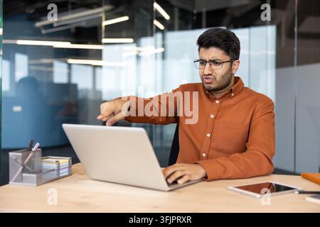 Frustrated businessman working on laptop in office, stressed employee pointing at screen with deadline pressure and professional burnout in a modern corporate workplace. Stock Photo
