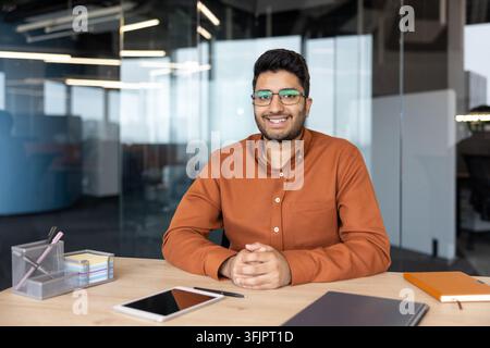 Professional businessman sitting at desk in modern office, smiling young man working with documents and tablet, corporate career and leadership concept for business success and workplace productivity. Stock Photo