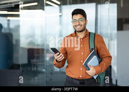 Young professional man holding a smartphone and documents in a modern office, smiling businessman with glasses and backpack ready for work, corporate career success and digital connectivity. Stock Photo