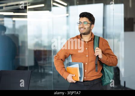 Young professional man with backpack and documents, smiling businessman in modern office environment for corporate career development and workplace success concept. Confident employee holding folders. Stock Photo