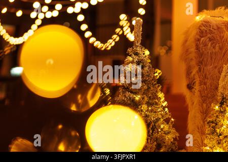 Close view of a frosted evergreen tree glowing with small lights next to large yellow bokeh highlights. Warm festive interior decoration during winter Stock Photo