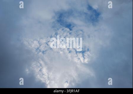 Soft white altocumulus clouds opening through dark overcast sky with patches of blue. Stock Photo