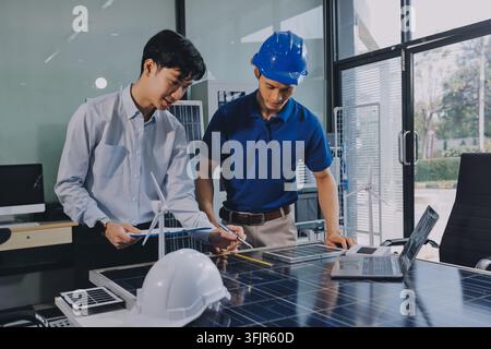 Two Asian engineers wearing hardhats and safety vests discussing solar panel installation and renewable energy project in the office. Stock Photo