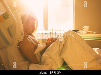 A young male patient on a bed in a ward uses a phone for fun Stock Photo