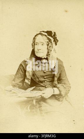 Photograph depicting Carte de visite of Anne Pratt, by William Clark (photographic business), from the Carte de Visite photography collection. A seated elderly woman wearing a dark dress with decorative buttons and lace cuffs, a lace head covering with ribbons, and a brooch at her collar, holding papers on a small table, in a sepia-toned photograph with beige and brown tones. 10.5 (h) x 6.3 (w) cm, Portrait, Photographic print. Printed inscription on recto, 'Clark Bristol'. Ink inscription on verso, 'For my dear Catherine'. Stock Photo