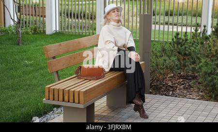 Older woman sits calmly on wooden bench in landscaped garden, eyes closed beneath white hat. Relaxed woman pauses beside brown handbag, finding Stock Photo