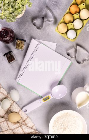 Flat lay showing baking ingredients like eggs, flour, milk, and spices arranged around an open blank notebook, with heart shaped cookie cutters ready Stock Photo