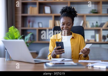 Financial management and remote work, woman using smartphone and holding receipt for expense tracking at desk with laptop in home office for business accounting and personal finance planning. Stock Photo