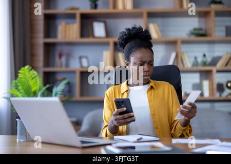 Financial stress and personal debt, woman checking bills and smartphone at office desk with laptop, worried person managing household budget and monthly expenses. Stock Photo
