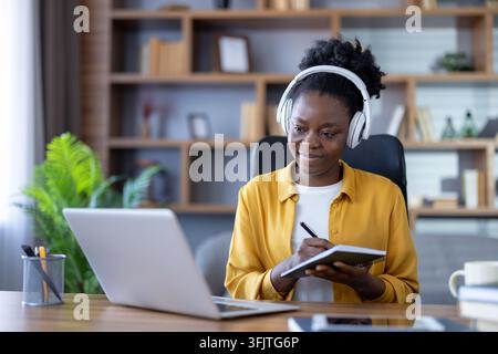 Remote work and online education, woman using laptop and headphones for virtual meeting or webinar while taking notes in a home office with a desk and bookshelf. Professional freelancer working. Stock Photo