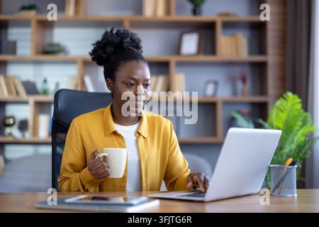 Remote work and digital nomad lifestyle, woman working on a laptop with coffee at a desk in a home office for professional business communication and productivity. Focused employee using computer. Stock Photo