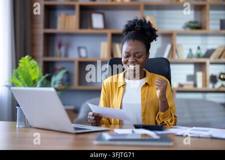 Successful businesswoman celebrating career achievement with documents and laptop in office. Happy professional woman reading good news and feeling excited at desk while reviewing important paperwork. Stock Photo