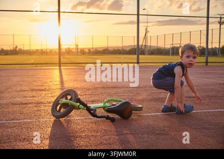 A toddler stands next to a fallen bicycle on an empty sports court during golden hour Stock Photo