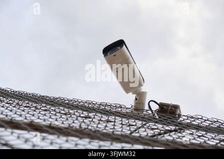 A white security camera mounted on a metal wire fence under an overcast sky background Stock Photo