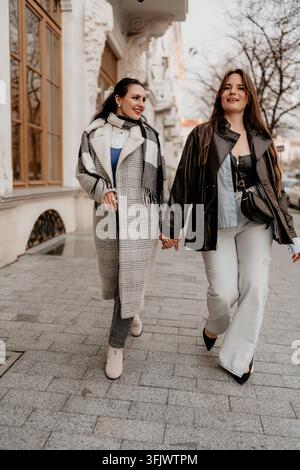 Women walking city strolling through urban streets while holding hands in stylish layered autumn fashion outfits and coats Stock Photo
