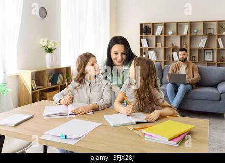 Mother helping two daughters with homework at home, father on laptop nearby Stock Photo