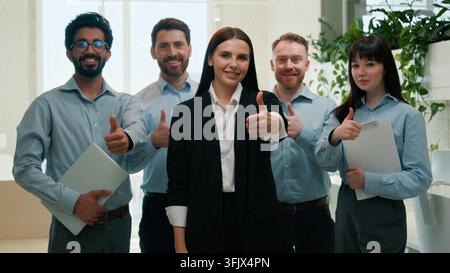 Happy multinational business team portrait showing thumbs up in modern office approval smiling businessmen businesswomen looking at camera colleagues Stock Photo