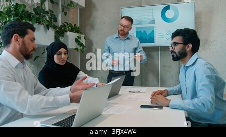 Man leader talking to multiracial coworkers business team indoors office conference employees businessmen muslim businesswoman in hijab discussing Stock Photo