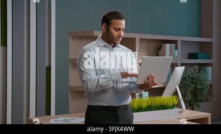 Happy Indian Arabian business man smiling looking at camera working laptop indoor office workplace corporate worker typing chatting online clients Stock Photo