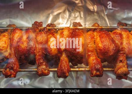 Street food roasted chicken legs on rotisserie with crispy browned skin, hot grilled poultry prepared for takeaway meal at food stall. Stock Photo