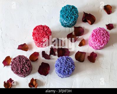 Five decorative rose-shaped candles in various vibrant colors are arranged on a white textured background with scattered dried rose petals. Stock Photo