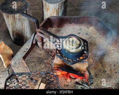 A close-up shot of a black kettle heating on burning logs inside a campfire pit at a national park in Lapland, Finland, surrounded by wooden seating s Stock Photo