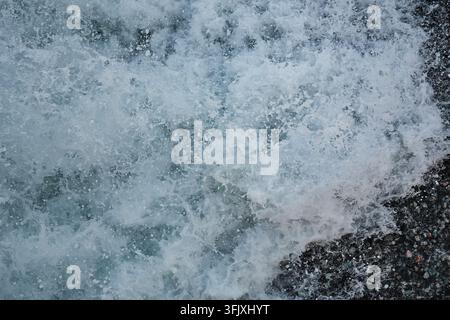 White sea foam and breaking water over a dark surface near the shore. Abstract natural water texture with a dynamic foamy pattern. Stock Photo