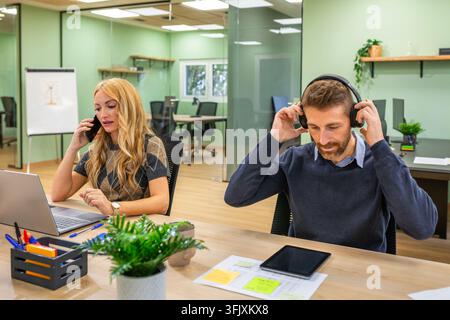 Professionals are working at their desks, a woman talking on the phone and using a laptop while a man is putting on headphones, showcasing productivit Stock Photo
