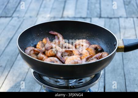 Delicious shrimps are frying in a black pan placed on a portable camping gas stove outdoors against a rustic wooden deck background, preparing a tasty Stock Photo