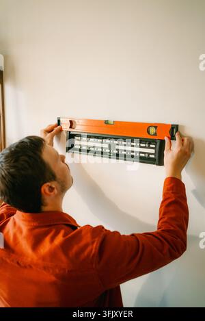 Man checking the level of a tv wall mount bracket on a white wall using a spirit level, demonstrating careful installation and diy accuracy for a home improvement project Stock Photo