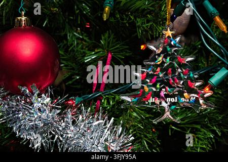 A close up view of holiday decorations hanging on a pine tree with glowing string lights and silver tinsel creating a warm seasonal atmosphere. Stock Photo