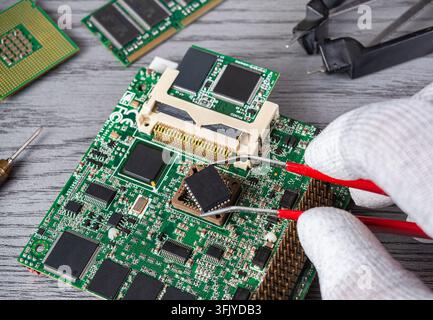 A technician handles a small chip on a circuit board using tweezers in a workshop filled with tools and equipment during the day. Stock Photo