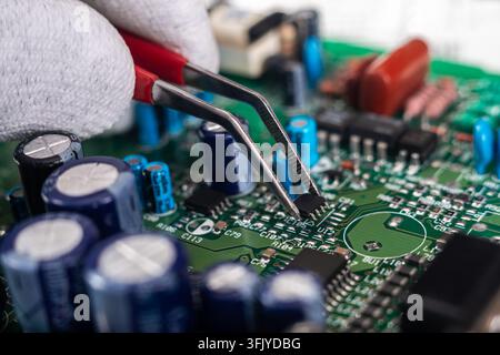 A technician uses tweezers to handle components on a circuit board in an electronics repair shop during daytime. Stock Photo