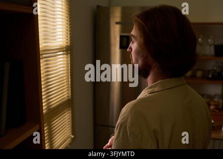 Thoughtful Man Looking through Window Blinds at Home Stock Photo