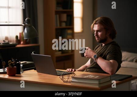 Man Drinking Coffee and Working on Laptop at Home Desk Stock Photo