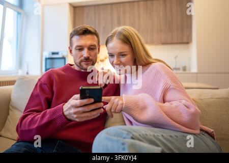 Happy married couple spending time together at home on sofa together scrolling on smartphone online. Stock Photo