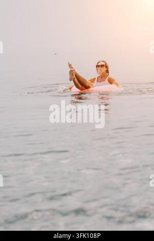 Woman inflatable ring ocean young woman relaxing on a pink donut tube in the calm sea during a warm sunset Stock Photo