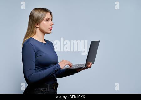 Portrait of a focused young blonde woman standing in profile and typing on a laptop. Woman in a dark blue long-sleeve top and black jeans in a studio Stock Photo