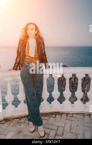Woman sunset coastal young lady leans on a white stone balcony railing enjoying a serene sea view during golden hour Stock Photo