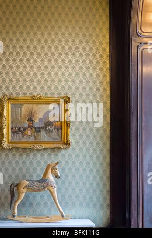 A carved wooden rocking horse stands on a marble console beneath a gilt framed painting of a rainy city street, set against patterned wallpaper and da Stock Photo