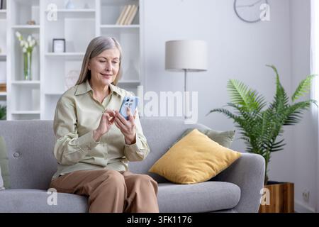 Mature woman with gray hair smiling and interacting with her mobile phone while relaxing comfortably on a modern couch in a bright domestic living room, embracing digital communication and leisure Stock Photo