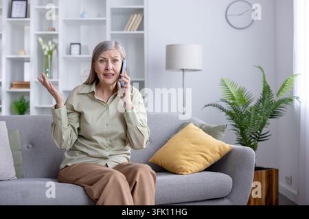 Senior woman sitting on a sofa in her living room, feeling frustrated and confused while having an angry conversation on her mobile phone, expressing disagreement and communication issues Stock Photo